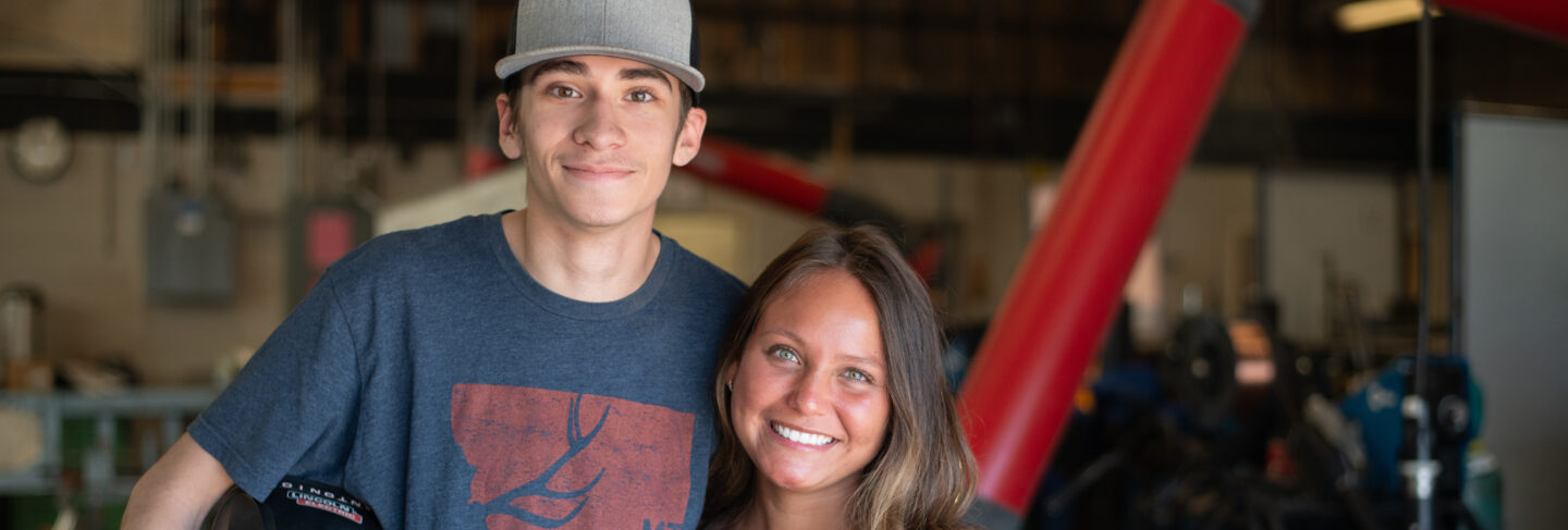 Two students smiling in a workshop at Flathead Valley Community College, one holding welding equipment.