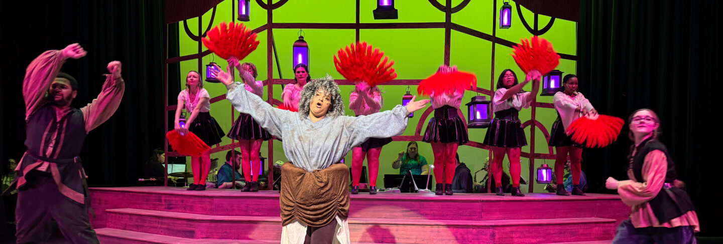 Actors performing on stage in a theatre production at Flathead Valley Community College, with a live orchestra in the background.