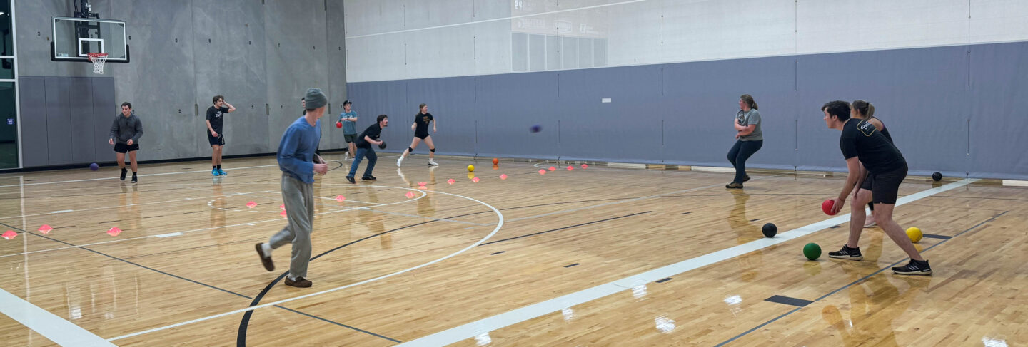 Students and staff playing a lively game of dodgeball in a campus gymnasium, with bright lighting and a polished wood floor.